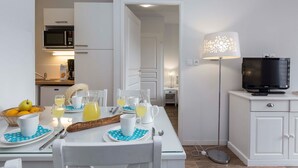 A dining area with a table set for breakfast, a microwave above the counter, a television on a cabinet, and a lamp with a decorative shade. - Studio cosy et charmant 2p (Hourtin)