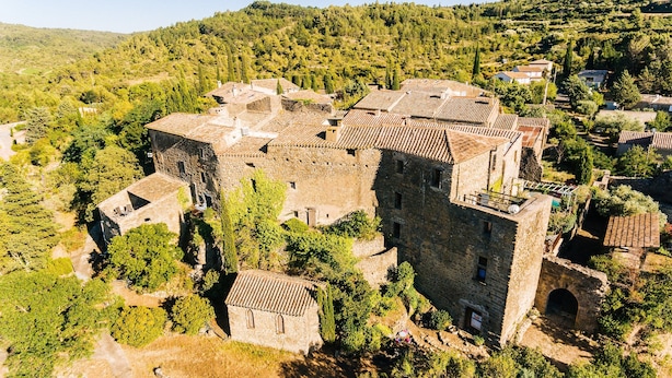 Vue aérienne d’un ensemble de bâtiments historiques en pierre, composé de plusieurs structures, entouré d’une végétation luxuriante et de collines.