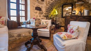 Une pièce chaleureuse aux murs de pierre, avec une table ronde en bois, un canapé blanc et un fauteuil agrémenté de coussins à motifs floraux.