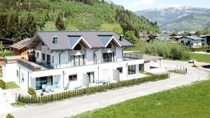 A modern two-story house with a balcony, surrounded by lush greenery and mountains in the background. - Finest Kitzblick Golf Suites TOP 2 (Zell am See)