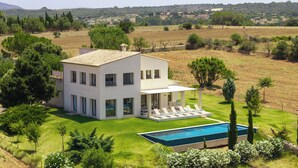 Une maison moderne à deux étages, dotée d’une piscine, entourée d’une végétation luxuriante et située dans un paysage vallonné.