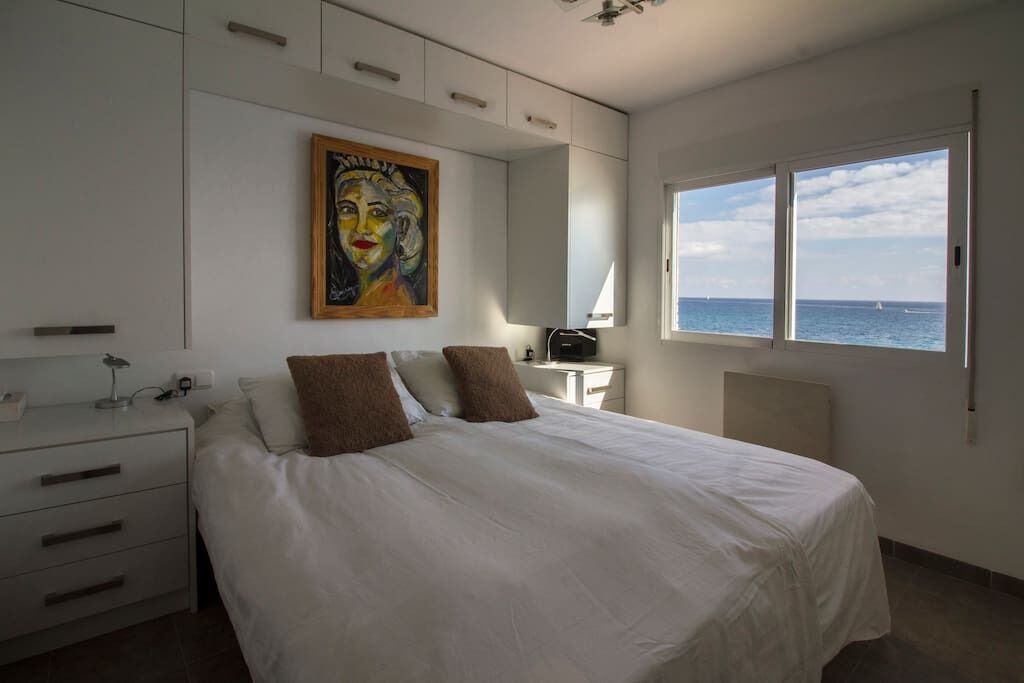 Un dormitorio con cama, mesita de noche, escritorio, silla, un cuadro y ventana con vista al mar.