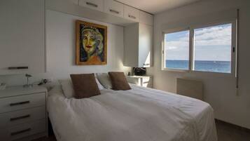Un dormitorio con cama, mesita de noche, escritorio, silla, un cuadro y ventana con vista al mar.