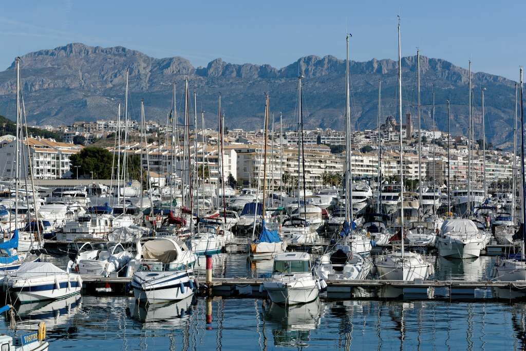 Una marina con numerosos barcos amarrados, rodeada de edificios y montañas al fondo.