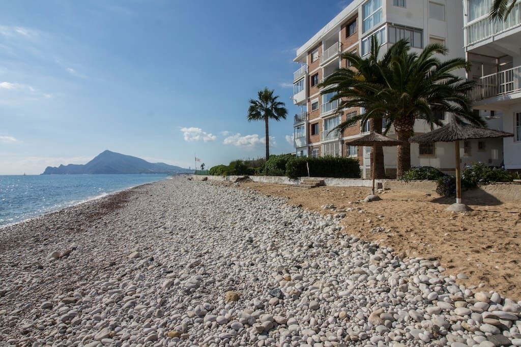 Una playa de guijarros con un edificio que tiene balcones y palmeras.
