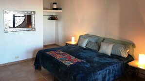 A bedroom with a large bed, a bedside table with a lamp, a wall-mounted mirror, and a shelf with clothes. - Spacious bright tiled 2 bedroom with Ocean View (Cerro)