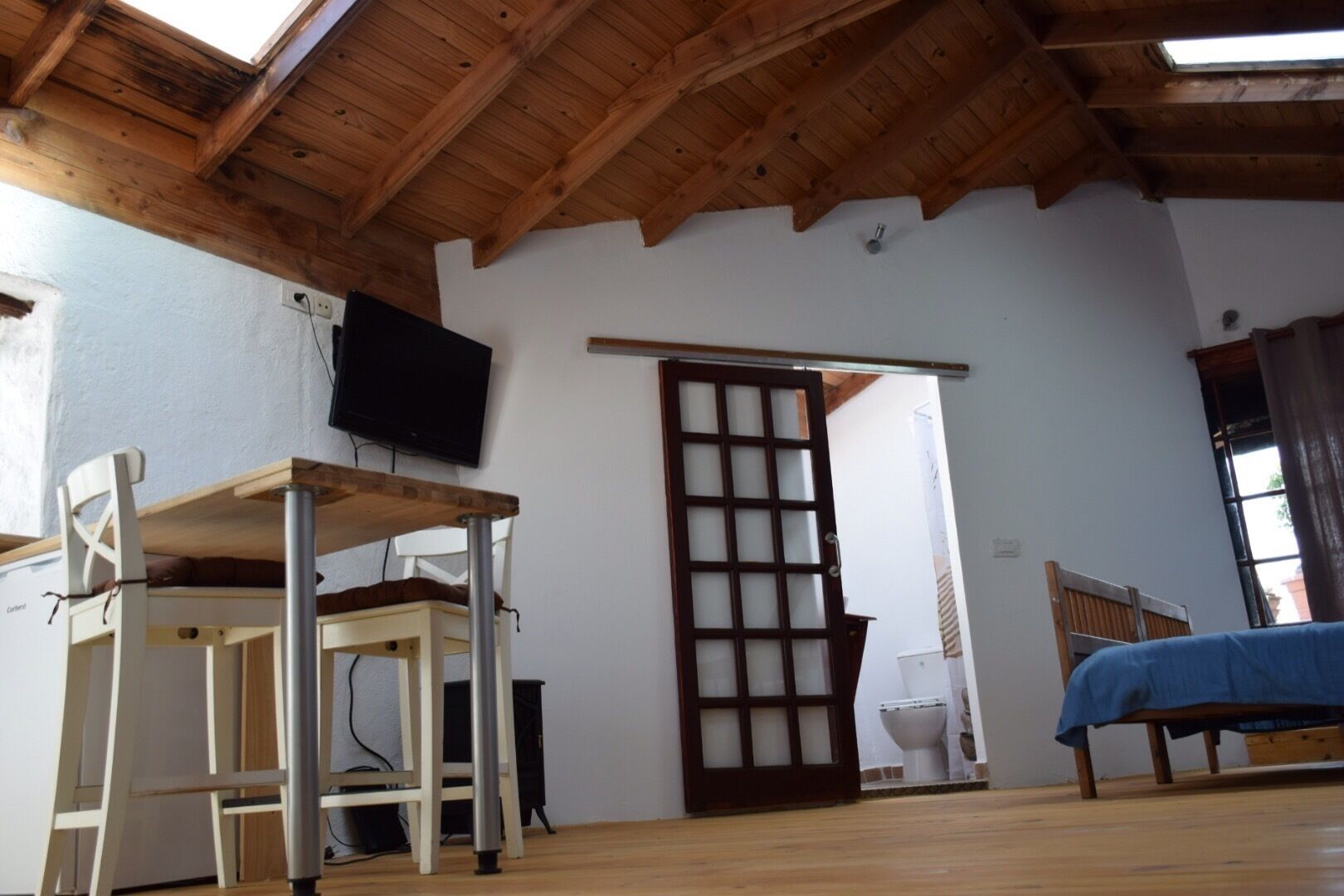 Une pièce avec un plafond en bois, un téléviseur fixé au mur, une porte en bois, un lit recouvert d’une couverture bleue et une petite table avec une chaise.