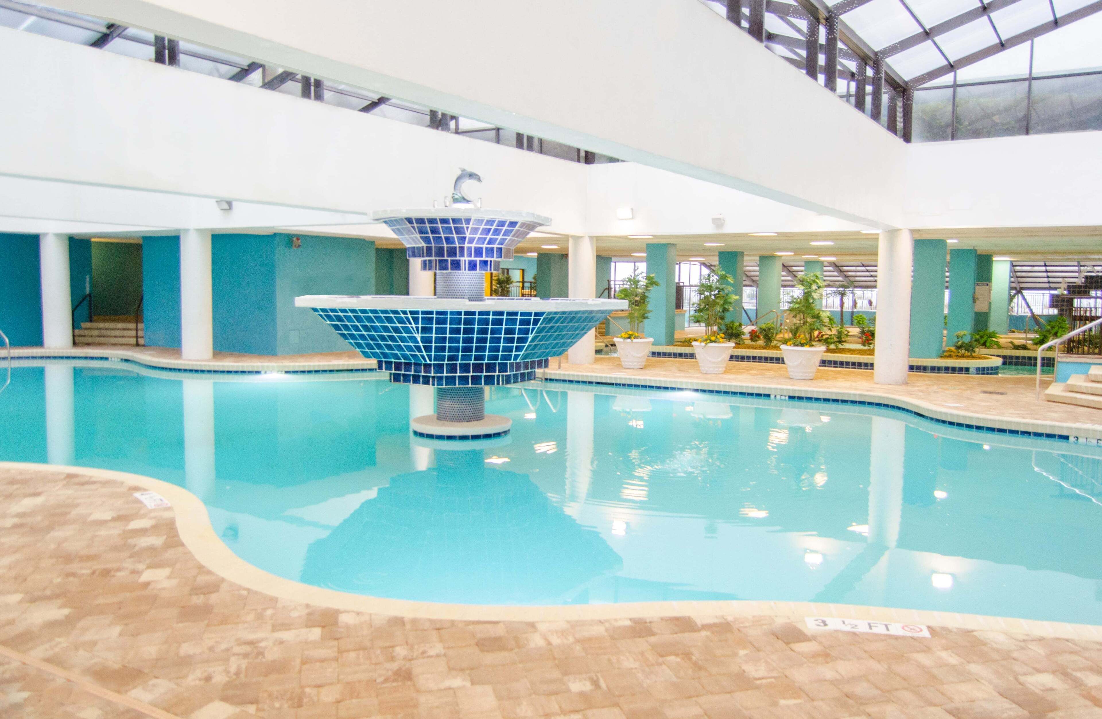 An indoor swimming pool with a central fountain, tiled walls, and potted plants around the perimeter.