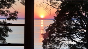 A sunset view through a window, with a tree in the foreground and a flat landscape in the distance. - Stunning Bayfront Views (Hampton Bays)