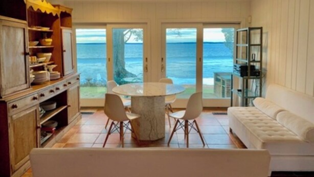 A dining area with a round table and chairs, a white sofa, and a view of the sea through large sliding doors. - Stunning Bayfront Views (Hampton Bays)