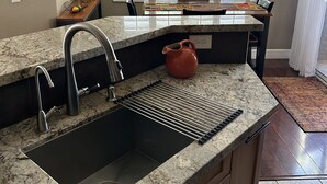 A kitchen with a granite countertop, stainless steel sink, and a built-in oven. There is a window with blinds, a potted plant, and a small table with a basket of fruit.