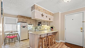 A kitchen with a central island, wooden stools, a dining table with chairs, and a white refrigerator.