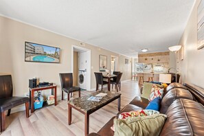 A modern living room with a sofa, coffee table, and chairs, a dining area with a table and chairs, a kitchen with a refrigerator and stove, and a laundry area with a washing machine.