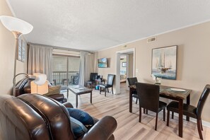 A living room with a leather sofa, a dining table with chairs, a TV mounted on the wall, and a balcony with a view.