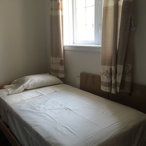 A single bed with white bedding, a window with curtains, and a plain wall.