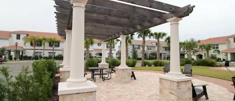 A covered patio area with stone columns and a pergola, outdoor seating, and a view of residential buildings in the background.