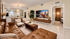 A modern living room with a large flat-screen TV, leather sofas, and a dining area in the background. - Sophisticated Condo, up to 6, Only 7 min to Disney (Kissimmee)