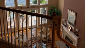 A spacious room with a large bay window overlooking a body of water, a ceiling fan, and a fireplace with a mantel. - Beautiful Saratoga Lake waterfront Townhouse + Community Amenities (Saratoga Springs)