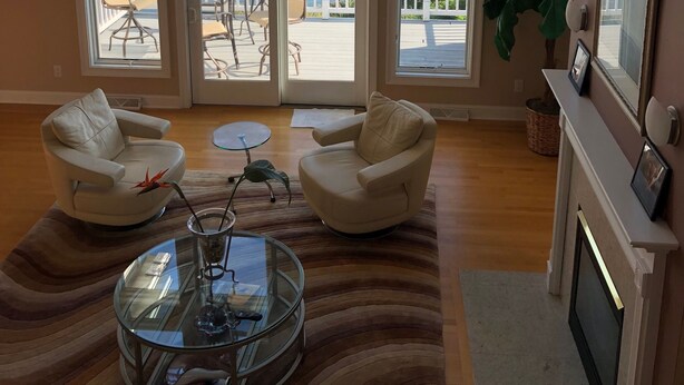 A spacious living room with large windows overlooking a body of water, two white armchairs, a glass coffee table, and a fireplace. - Beautiful Saratoga Lake waterfront Townhouse + Community Amenities (Saratoga Springs)