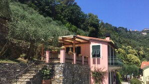 Una villa rosa con muro de piedra, escalera de madera y balcón con vistas a una colina.