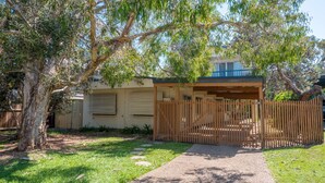 A covered outdoor area with a wooden fence, a pathway leading to a building with a blue roof, and a large tree providing shade. - Ngungun Street 10, Dicky Beach (DICKY BEACH)
