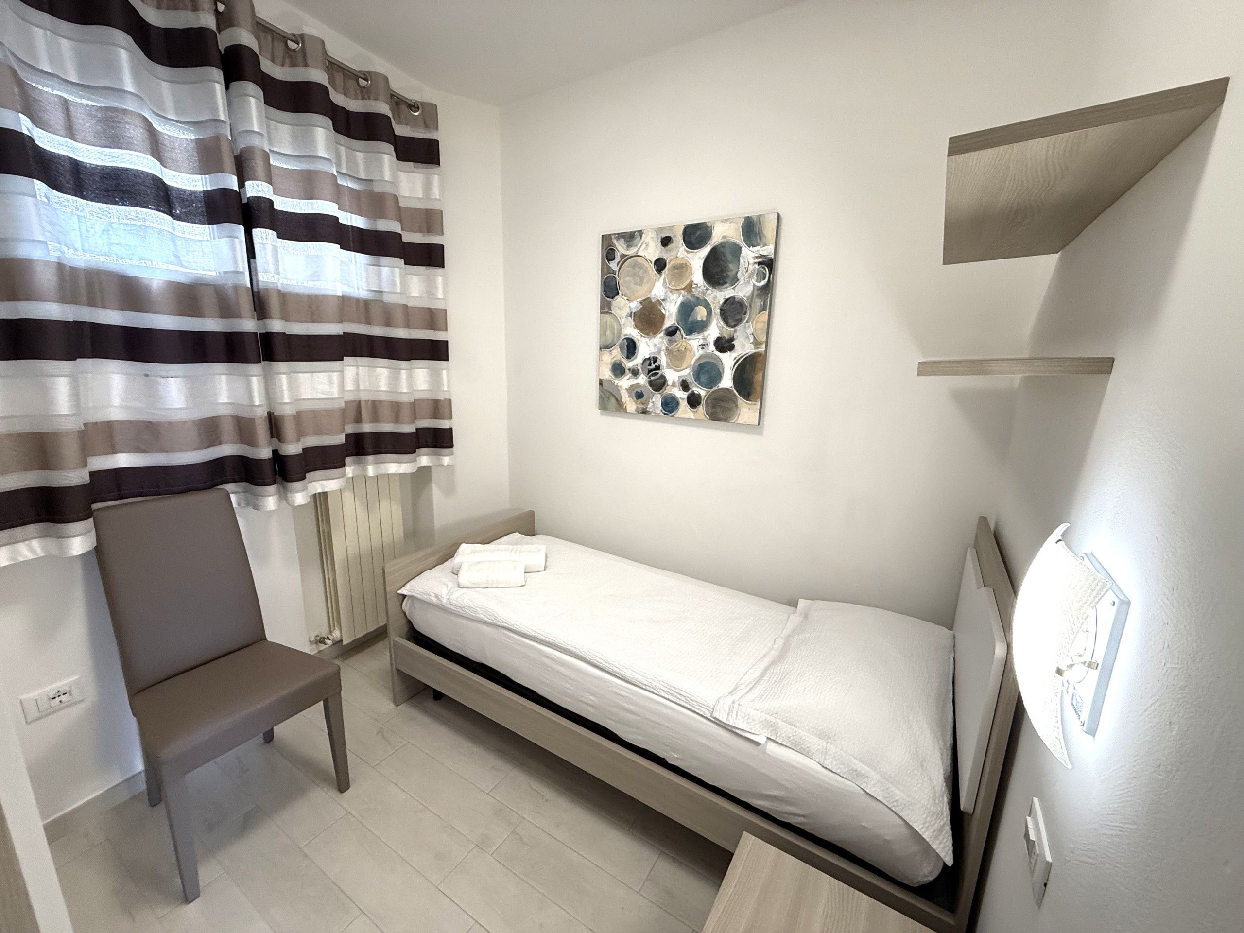 A single bed with a wooden headboard, a bench, a striped curtain, and a wall-mounted abstract art piece.