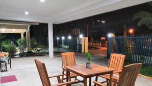Un patio al aire libre con una mesa y sillas de madera, rodeado de plantas en macetas y un piso de concreto.