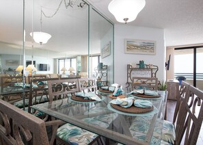 Un coin repas avec une table en verre, des chaises dotées de coussins à motifs et un grand miroir qui reflète la pièce.