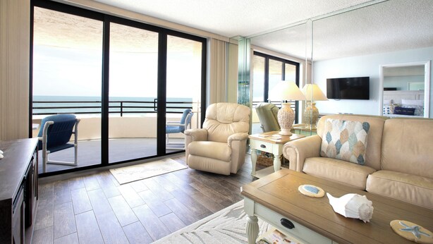 Une chambre d’hôtel avec un balcon, un canapé, un fauteuil, une télévision et un bureau.