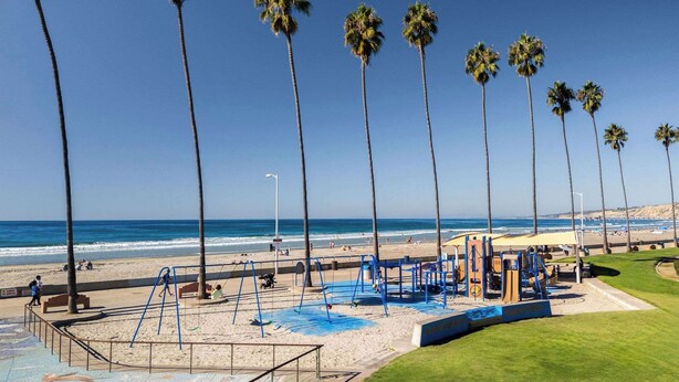 A beach playground with swings and slides, palm trees, and a sandy beach. - Beach Bungalow 3. Dog-Friendly. Steps from the beach! (San Diego)
