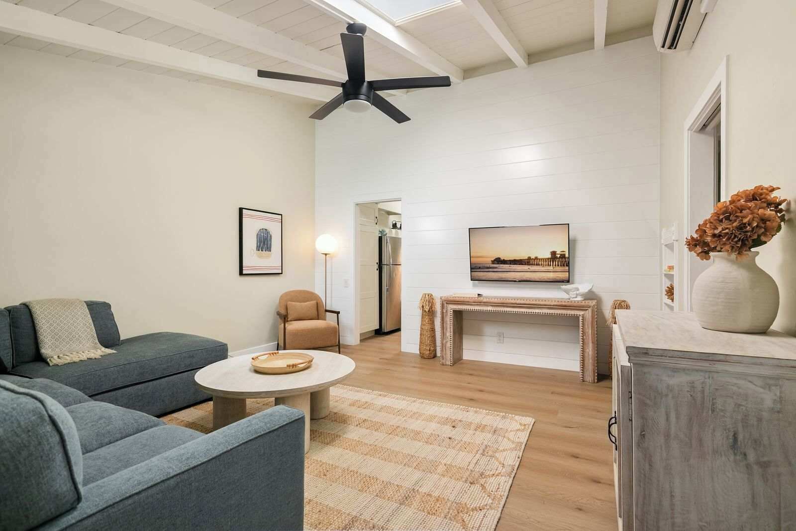 A modern living room with a grey sofa, a round coffee table, a beige armchair, a TV on a wooden stand, and a ceiling fan.