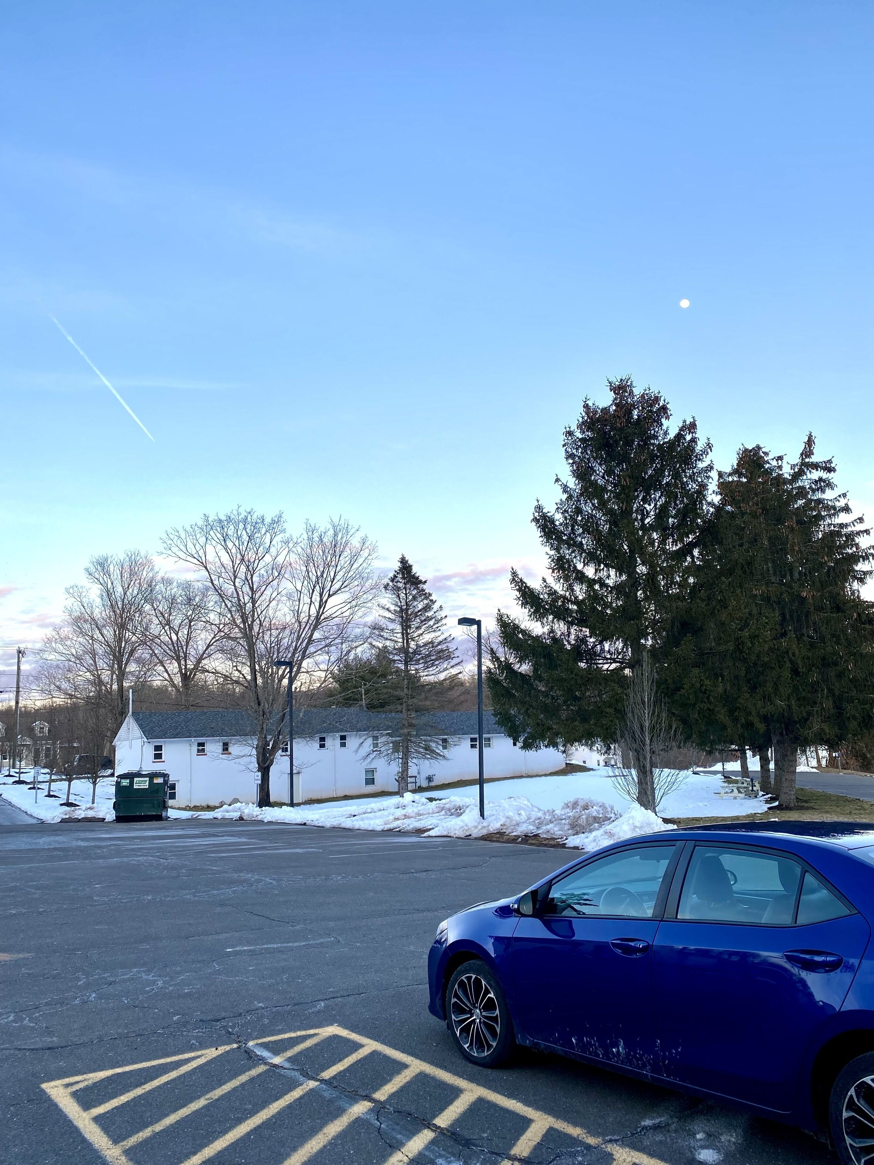A parking lot with a blue car, snow on the ground, and trees in the background.