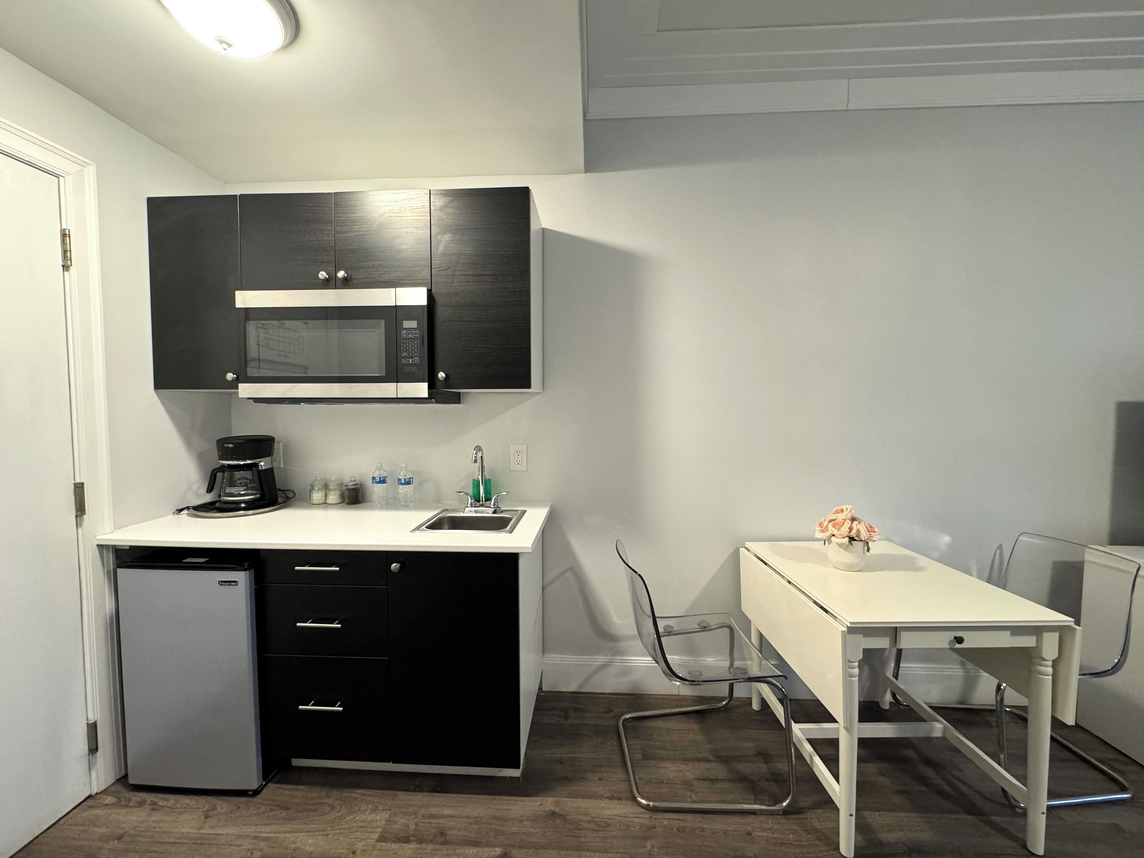 A compact kitchen with a microwave, coffee maker, and a small refrigerator. There is a white table with a vase of flowers and a chair.