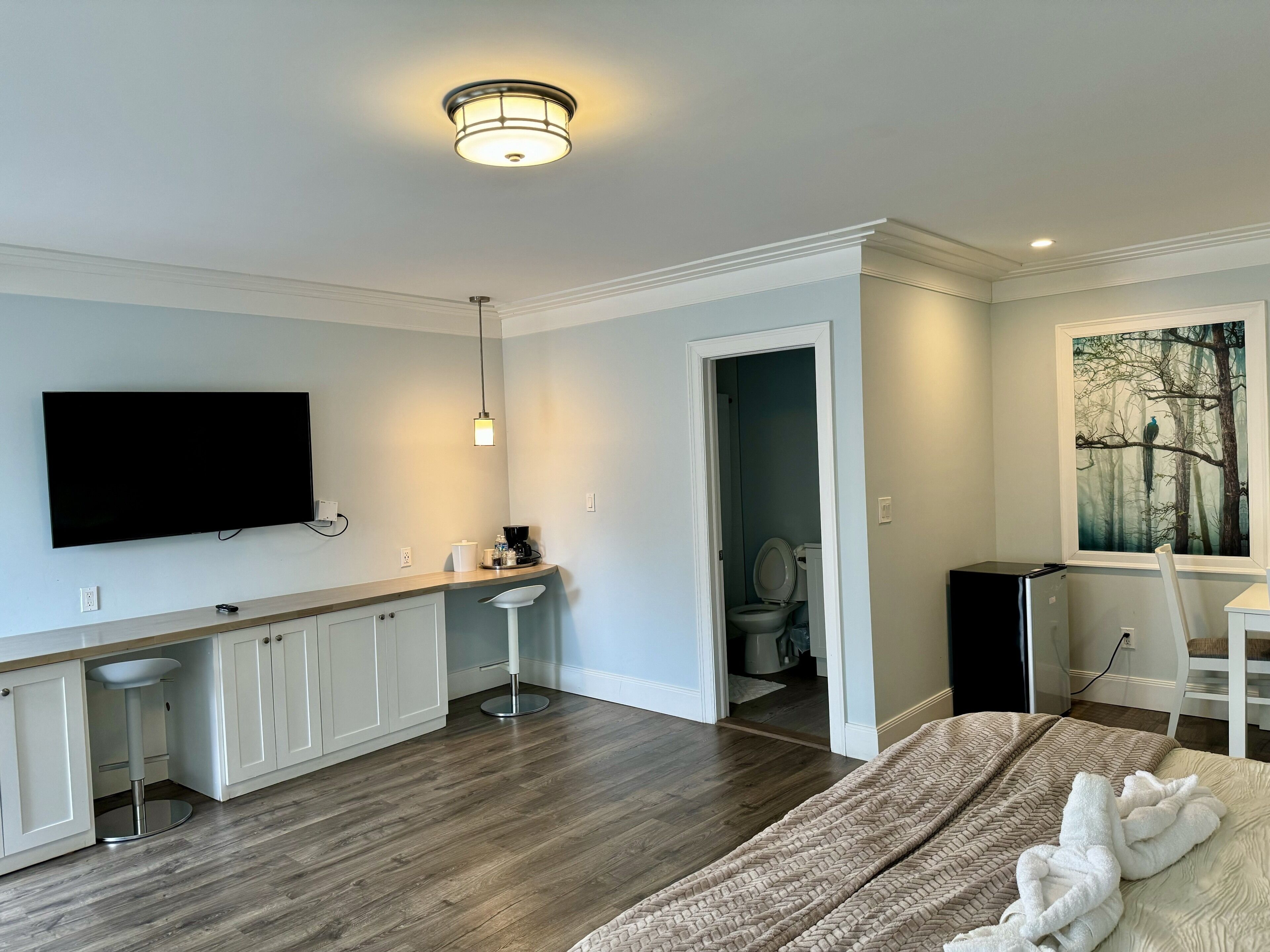 A modern hotel room with a flat-screen TV, a kitchenette, a large bed, and a painting on the wall.