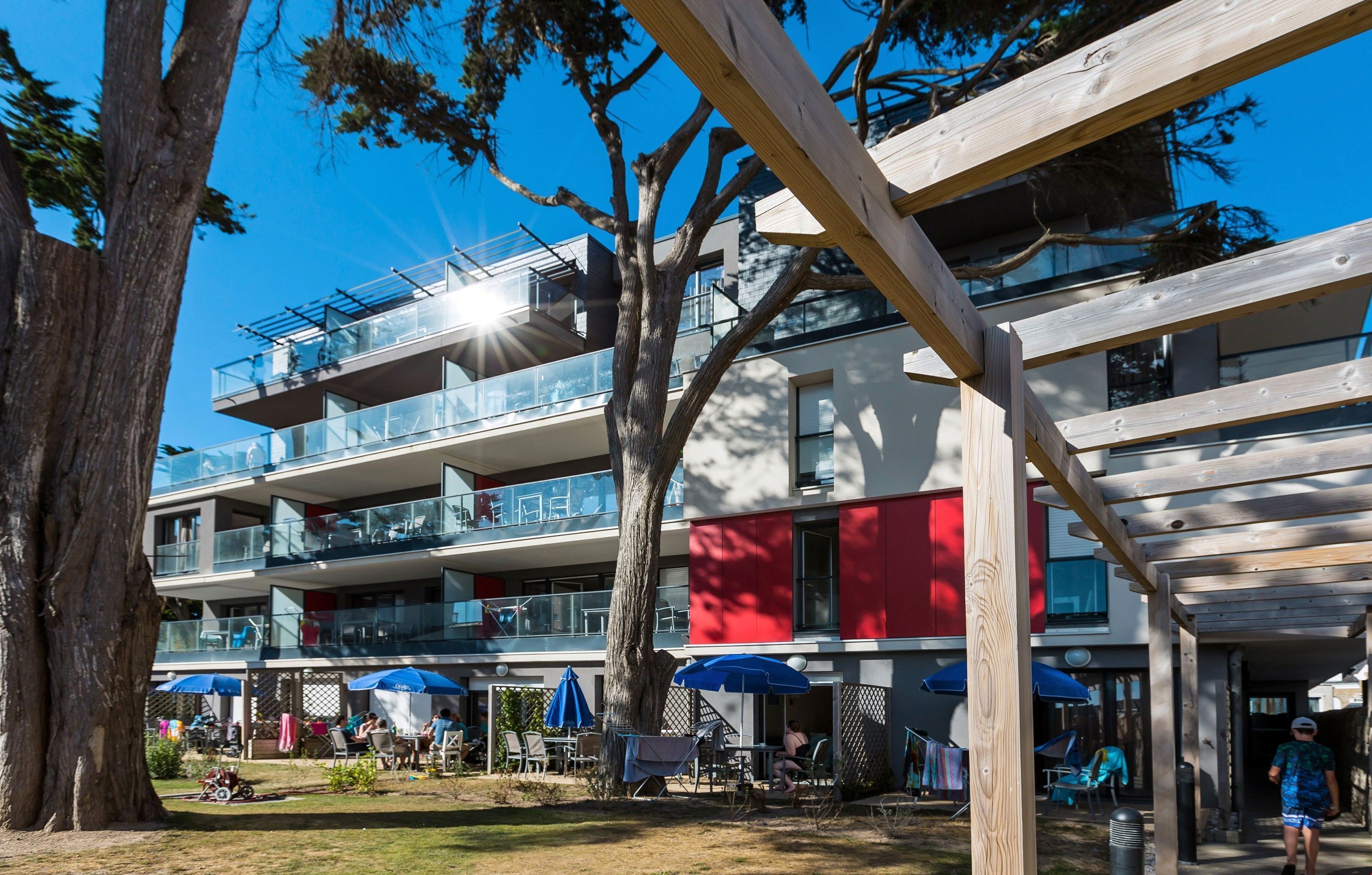 Un immeuble à plusieurs étages doté d’espaces extérieurs aménagés pour s’asseoir, d’une terrasse couverte et offrant une vue sur les arbres environnants.