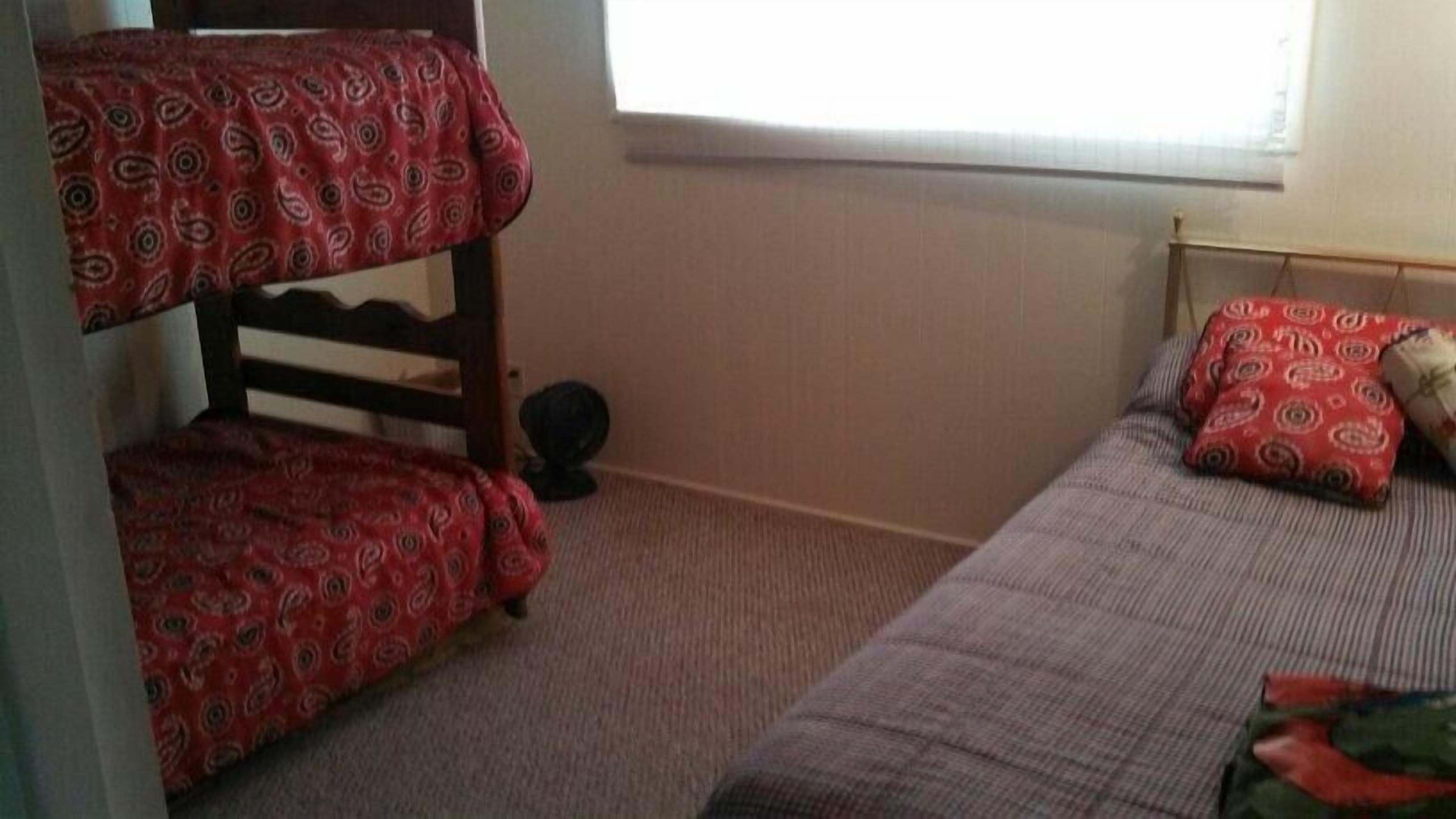 A bunk bed with a red patterned bedspread, a single bed with a grey blanket, and a window with white blinds.