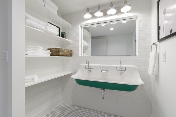 Un baño con un lavamanos blanco, lavabo verde y paredes de azulejos blancos. Se observan estantes con toallas y una cesta de mimbre.
