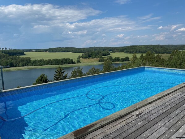 A clear blue swimming pool with a wooden deck, surrounded by a lake and forested hills. - Big House With amazing sea view (Grödinge)