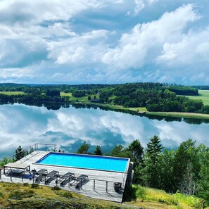 Vista chiara di una piscina con lettini da relax e un pontile in legno, immersa nel verde lussureggiante e affacciata su un lago tranquillo.