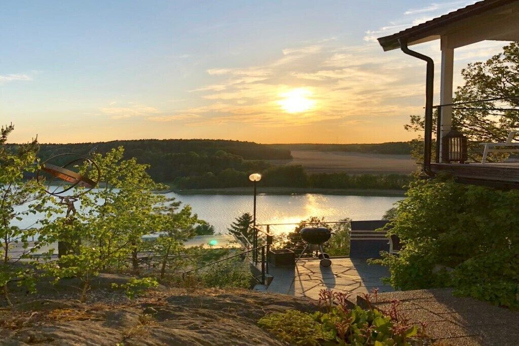 Un patio junto al lago con una parrilla, una lámpara y vistas al lago y colinas boscosas al atardecer.