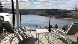 Un balcon avec des chaises blanches et une petite table, d’où l’on peut admirer un lac et des collines boisées.