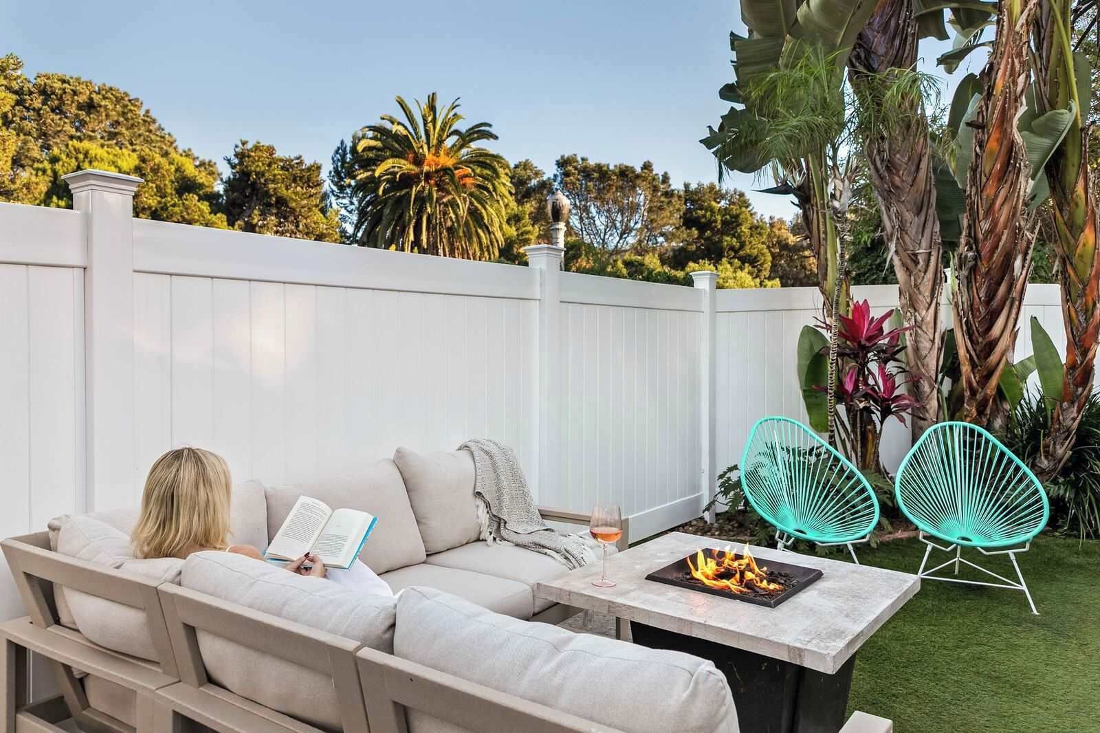 An outdoor seating area with a sofa, two chairs, a table with a fire feature, and a white fence.