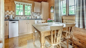 A wooden cabin with a kitchen featuring a dishwasher, a dining table with chairs, and a window with curtains.