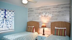 Quarto com duas camas, uma janela com persianas, uma luminária na parede e um ventilador de teto.