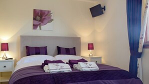 A neatly made bed with purple bedding, two bedside lamps, a flat-screen TV mounted on the wall, and a floral painting above the bed. - 2 bedroom accommodation in Auchterarder (Auchterarder)