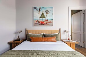 Un dormitorio con cama, almohadas, mesita de noche, lámpara, un cuadro en la pared y una puerta.