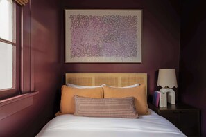 Un dormitorio con cama, cabecero, una obra de arte en la pared, mesita de noche con lámpara y ventana.