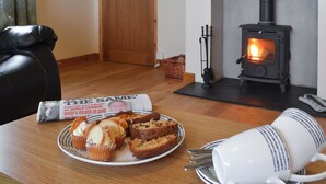 A cozy room with a wood-burning stove, a leather sofa, a dining table with a plate of muffins, and a newspaper.