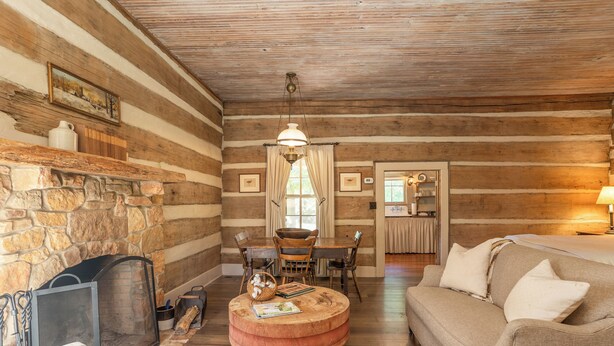 Interior de una cabaña de troncos con chimenea de piedra, mesa de comedor de madera y zona de estar acogedora.