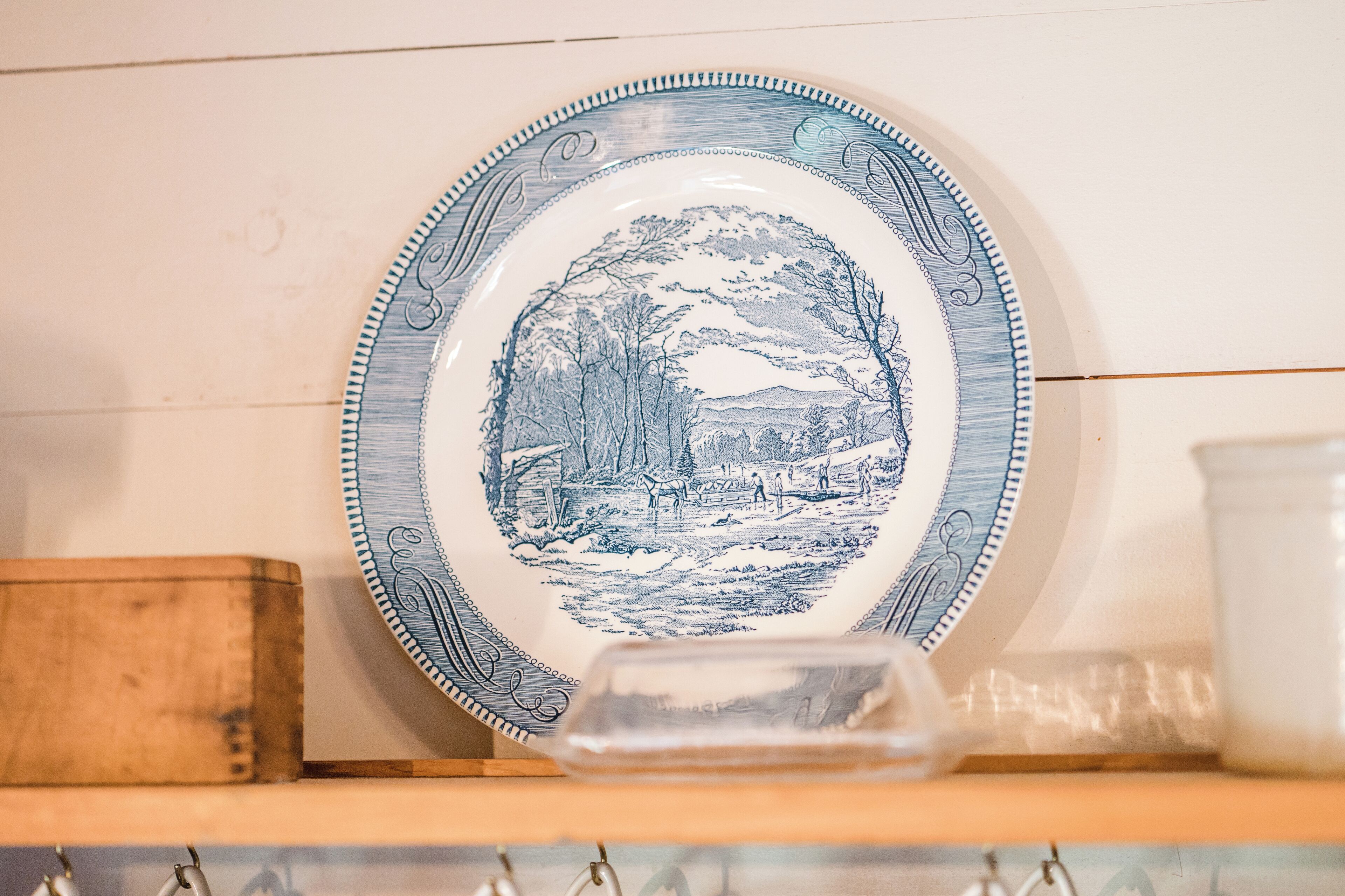 Une assiette en céramique ornée d’un motif de paysage bleu, posée sur une étagère en bois.